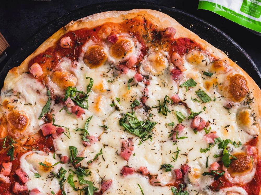 En pizza med ost, kjøttbiter og grønne urter på en svart pizzabunn, med en grønn brus i bakgrunnen.