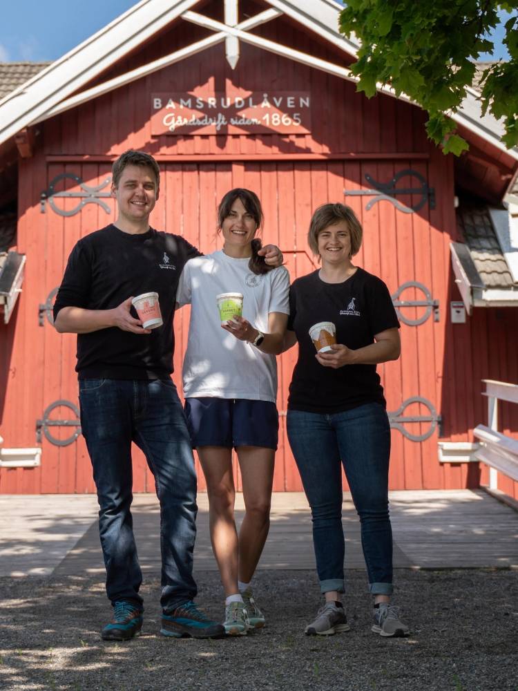 Tre personer står foran en rød bygning med skilt som sier Bamsrudlåven, og holder iskremer i hendene.