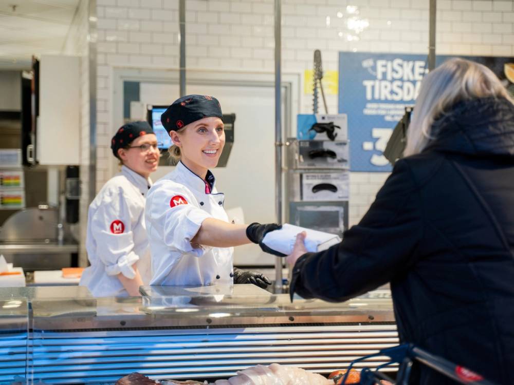 Kokk i hvit uniform og to assistenter i kjøkkenet som serverer en kunde i en matbutikk eller restaurant, med menyen på veggen i bakgrunnen.