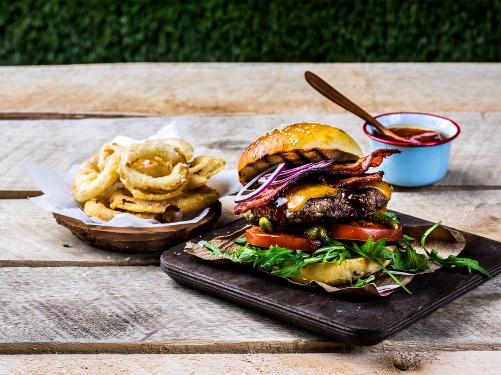 En burger med bacon, salat, tomat og løk på en svart skjærefjøl, med en liten skål med saus ved siden av.