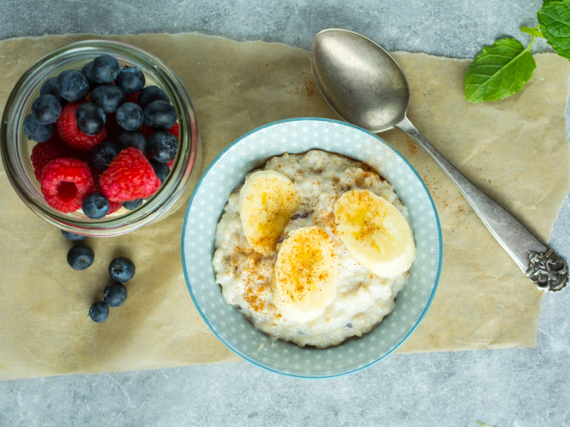 En skål med blåbær og bringebær, en skål med havregrøt toppet med bananskiver og honning, og en skje på et fat med grønne blader ved siden av, på et kjøkkenbord.