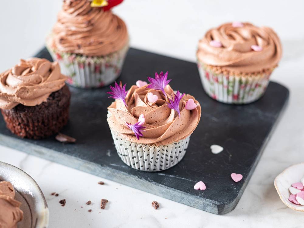 Fire cupcakes med sjokoladefrosting og små hjerte- og blomsterdekorasjoner på en svart steinplate.
