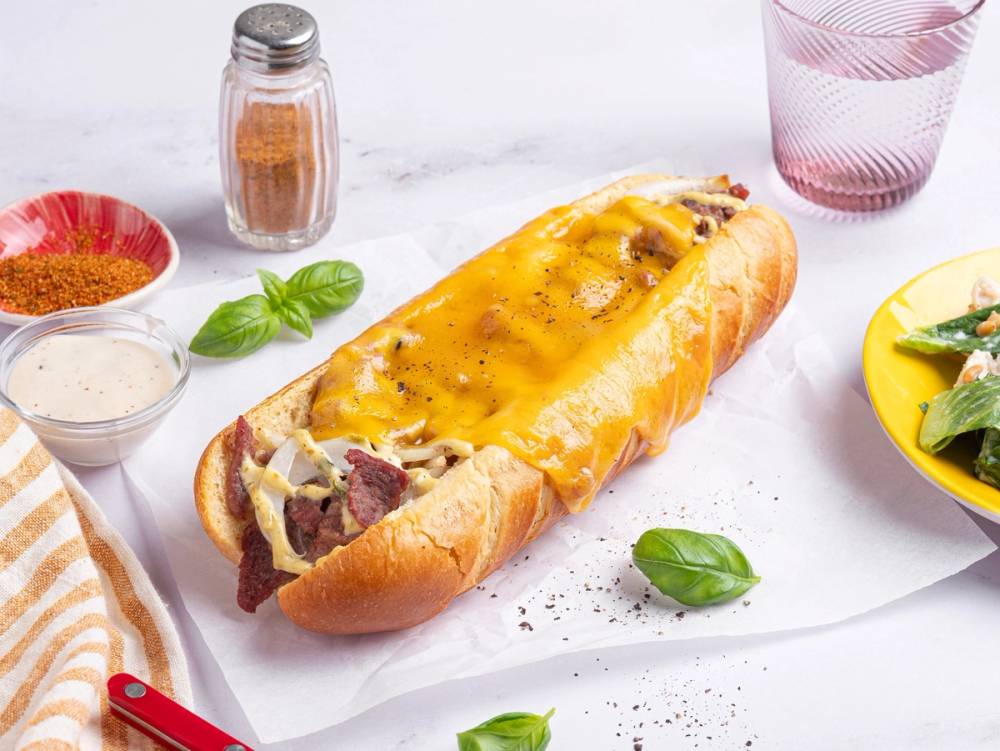 En baguette fylt med kjøtt og smeltet ost, omgitt av krydder, urter og tilbehør på et hvitt bord.