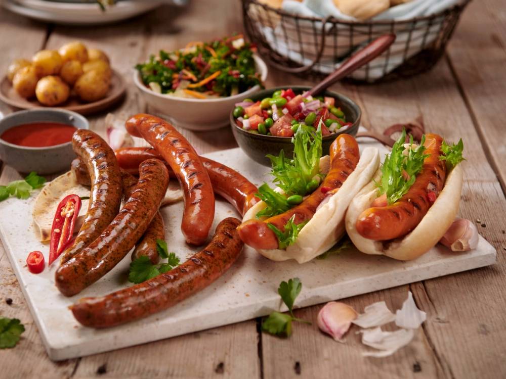Grillpølser med grønnsaker og tilbehør på et bord, inkludert en bolle med salat og en liten skål med ketchup.
