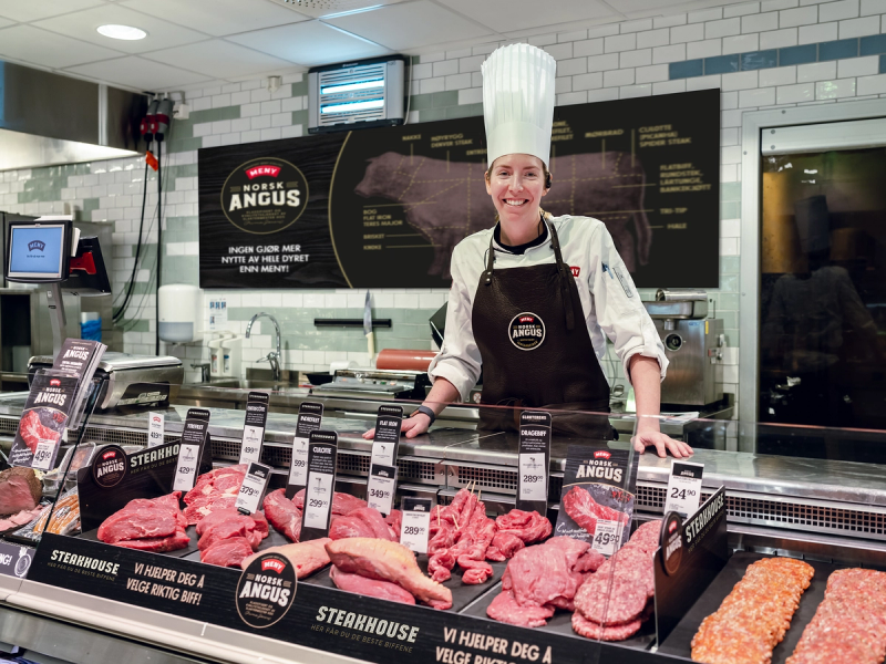 Kjøttvarer og Angus-merkede produkter i en kjøttdisk på en butikk, med en person bak disken som har ansiktet dekket av en stor skygge.