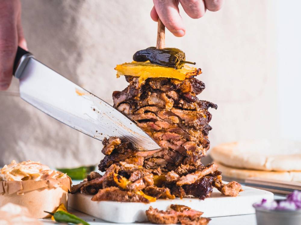 En person skjærer opp en stor mengde shawarma med en stor kniv, med kjøttstykker stablet opp på en tallerken.