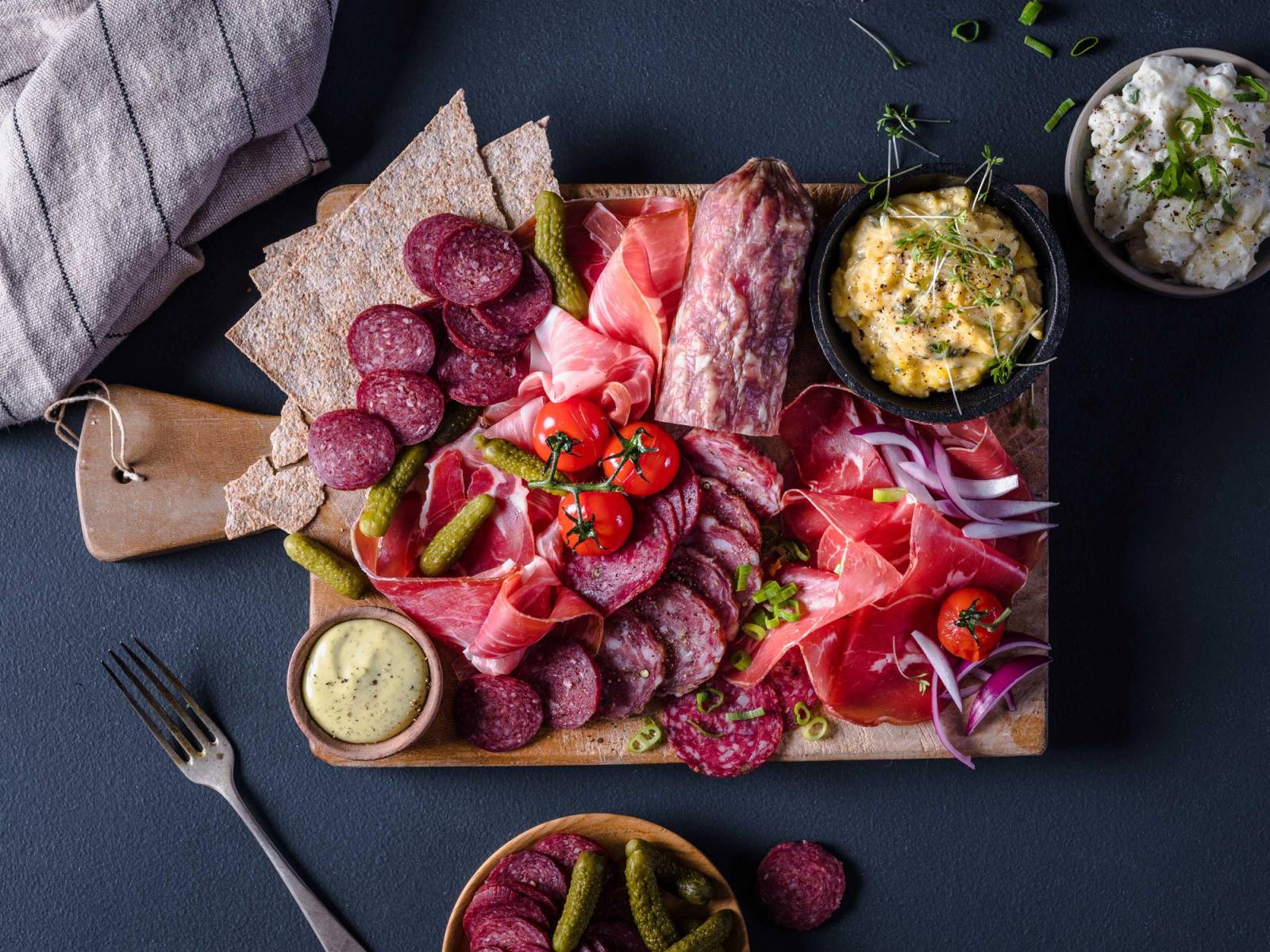 Et fat med ulike typer spekemat, pølser, og tilbehør som ost og urter, på en mørk bakgrunn.