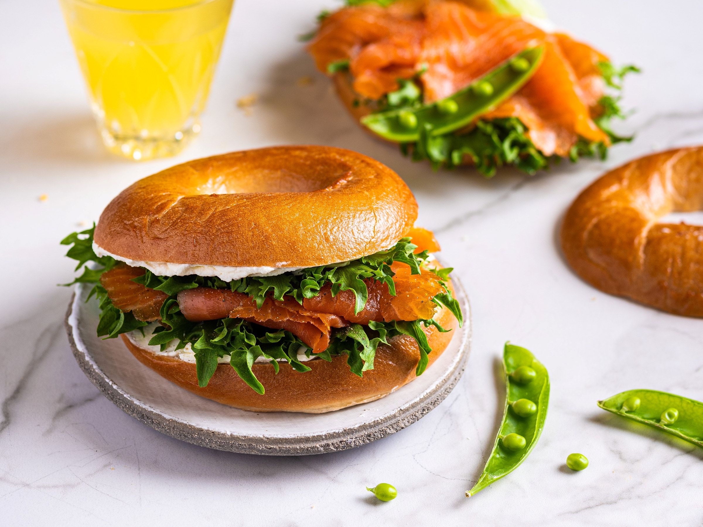 Bagel med røkelaks, kremost og salat på en tallerken, med en glass sitronvann i bakgrunnen.