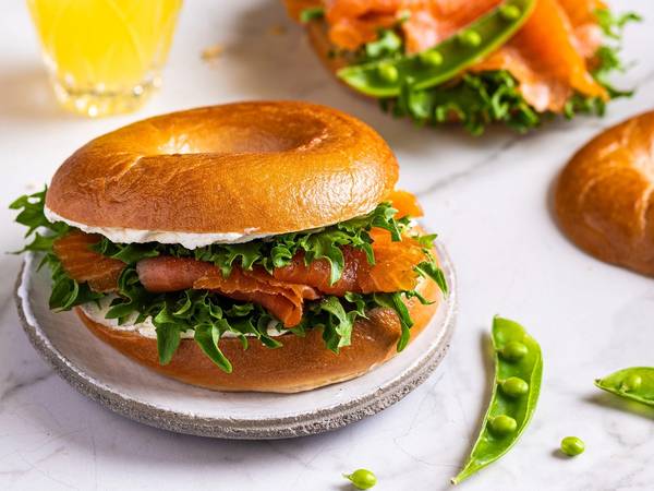 Bagel med røkelaks, kremost og salat på en tallerken, med en glass sitronvann i bakgrunnen.