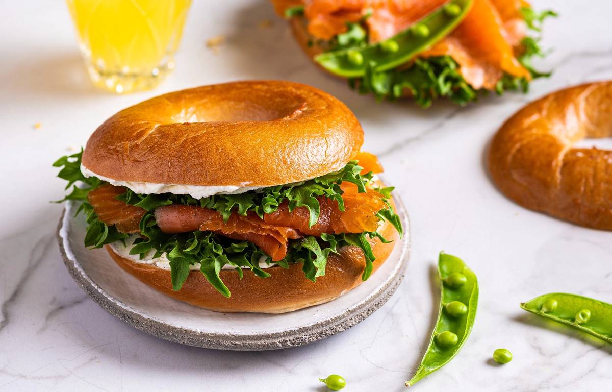 Bagel med røkelaks, kremost og salat på en tallerken, med en glass sitronvann i bakgrunnen.