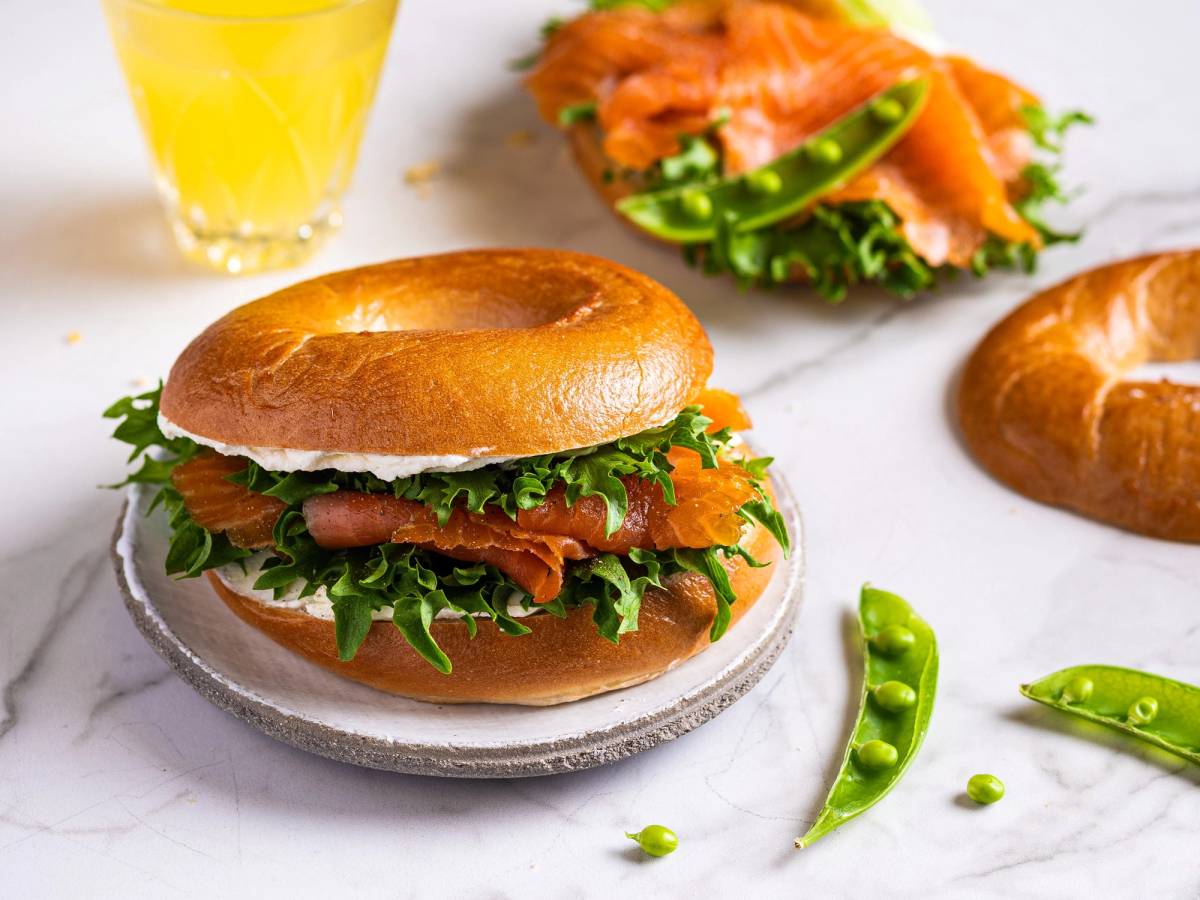 Bagel med røkelaks, kremost og salat på en tallerken, med en glass sitronvann i bakgrunnen.