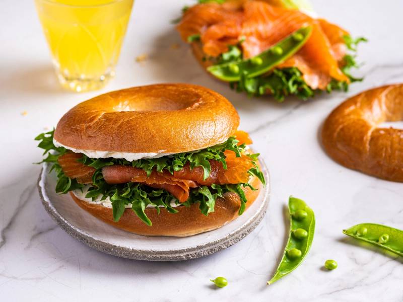 Bagel med røkelaks, kremost og salat på en tallerken, med en glass sitronvann i bakgrunnen.