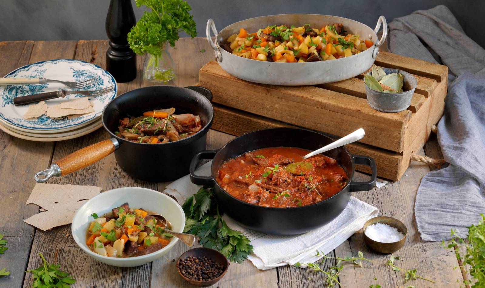 En rekke gryter og kasseroller med ulike retter står på et kjøkkenbord, inkludert en tomatsaus, en grønnsaksgryte og en gryte med kjøtt, omgitt av serveringsutstyr og urter.