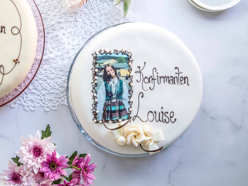 En konfirmasjonskake med bilde av en jente i konfirmasjonsantrekk og teksten 'Konfirmanten Louise' skrevet på kaken, omgitt av blomster og servietter.