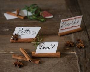 Små navnelapper med navnene Petter, Lene og Henrik, omgitt av kanelstenger, stjerneanis, og grønn urt på et treverk.