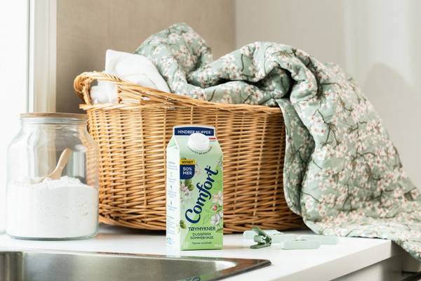 En kurv med sengetøy og en kartong med Comfort-vaskemiddel på et bord, med en liten del av et kjøkken i bakgrunnen.