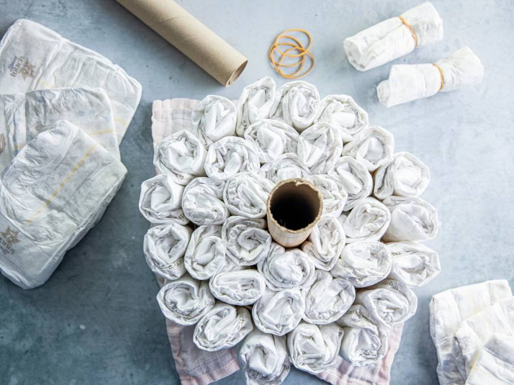 En bleiekake laget av sammenrullede bleier, dekorert med en liten papirkopp i midten, omgitt av flere bleier og annet babyutstyr på et lyst bord.