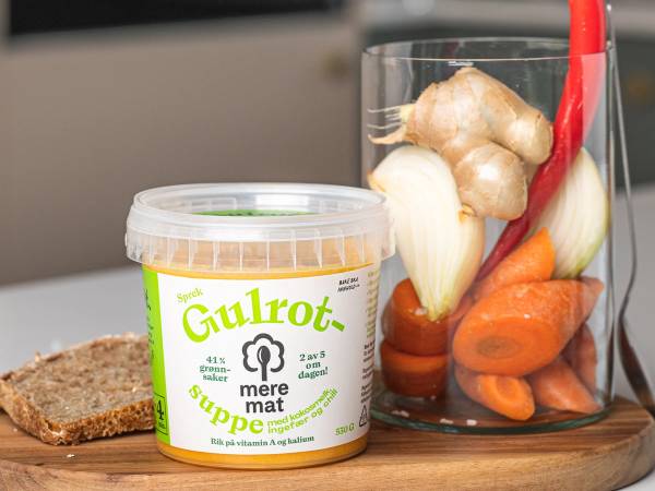 En glass med gulrot, løk og ingefær ved siden av en skål med gulrotsuppe, alt plassert på et kjøkkenbord.