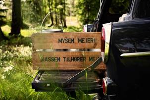 En melkekasse med melk fra nabogårdene står i bakluken på en bil i en grønn skogkledd natur.