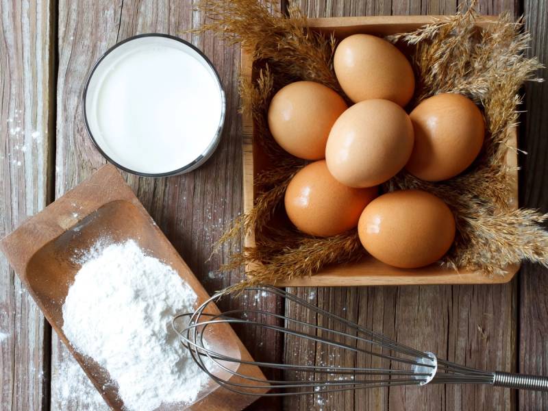 Seks egg i en kurv, en skål med melk og en skål med mel på et trebrett, brukt i matlaging.