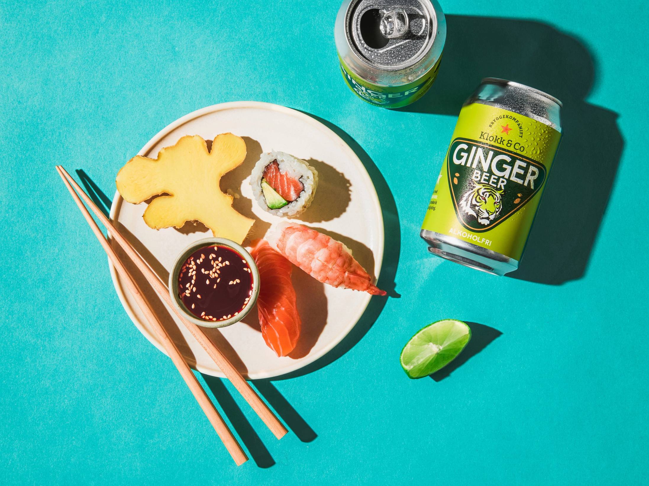 Sushi med laks, en bit med jordbær og en liten skål med soyasaus på en hvit tallerken, sammen med en kanne med ginger beer og en grønn lime på en blå bakgrunn.