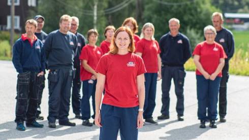 En gruppe ansatte ved Dovre Ysteri står ute på en parkeringsplass, alle iført røde og blå arbeidsklær, med en person foran som ser ut til å lede eller presentere.
