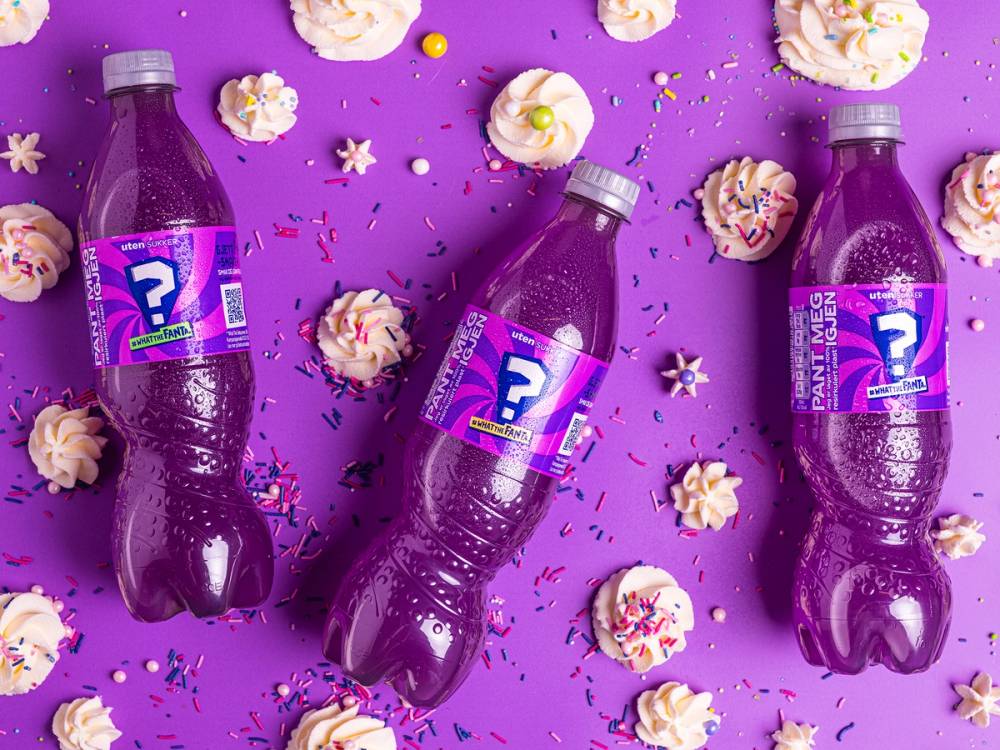 Tre flasker med lilla Fanta plassert på en lilla bakgrunn med små kaker og konfetti rundt.