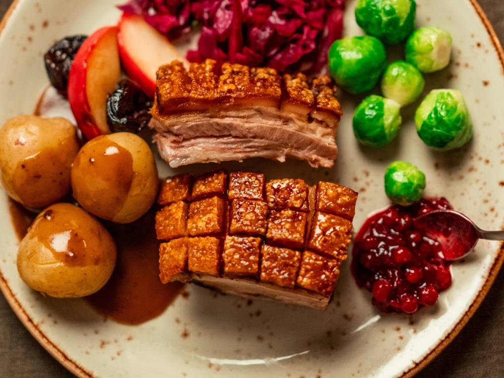 Stekt svinekjøtt med sprø svor, servert med poteter, grønne erter og tyttebærsyltetøy på en tallerken.