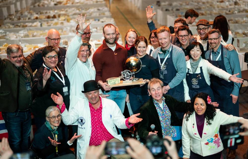 En gruppe mennesker samles rundt en person som holder en pokal og en liten modell av en bil, under en utendørs arrangement.