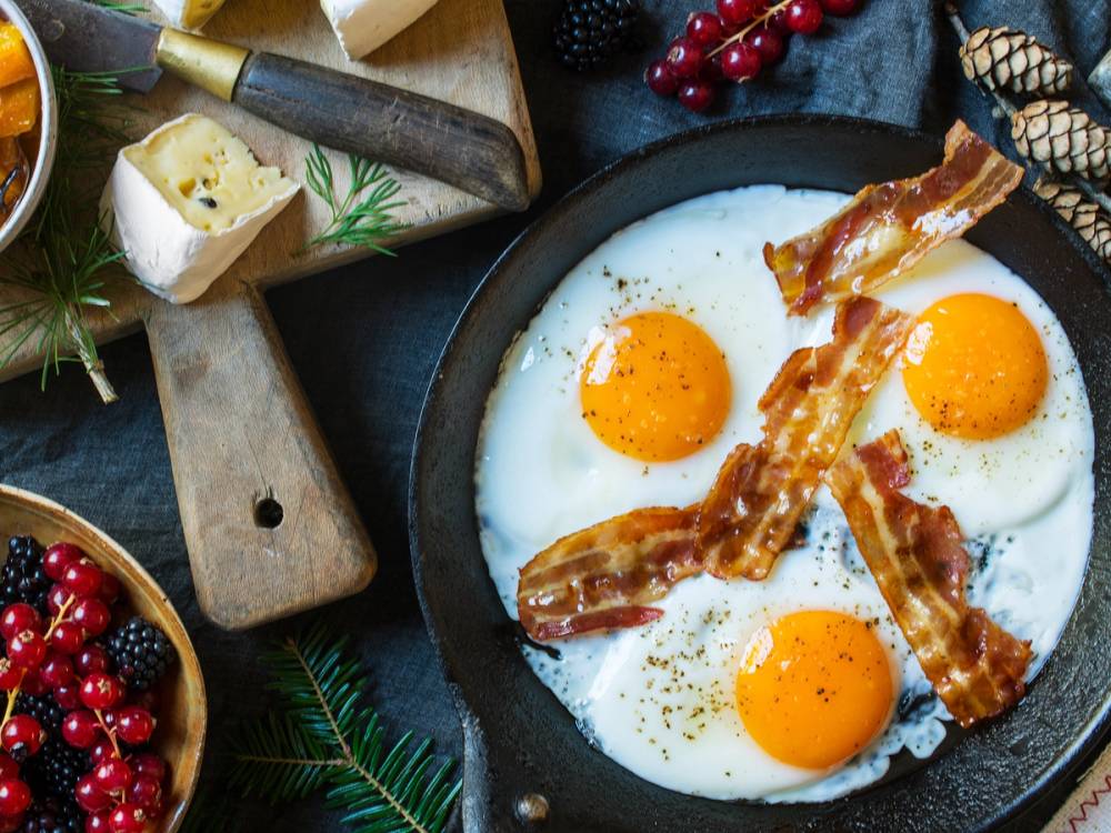 Stekte egg med sprøtt bacon på en svart tallerken, omgitt av bær og urter.