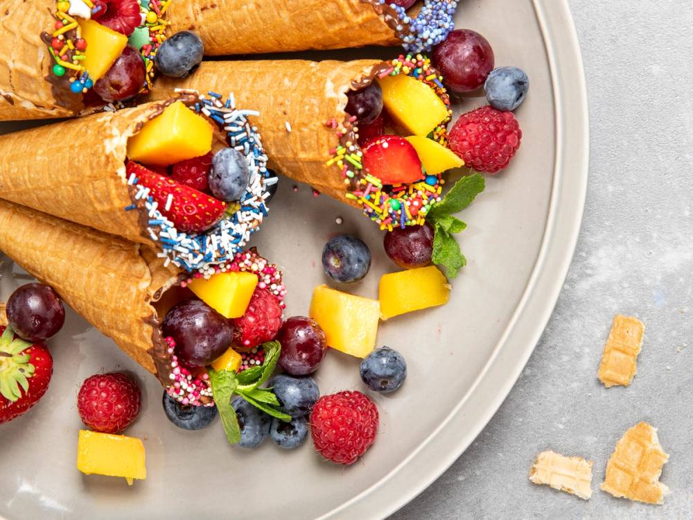 Tre isoppskåler fylt med bær og frukt, pyntet med fargerikt sukker og små grønne blader, på en hvit tallerken.