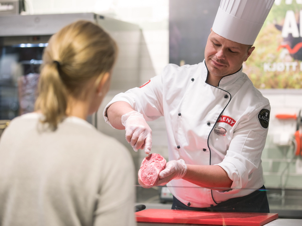 En person i kokkeforkle viser et stykke kjøtt til en kunde i en butikk eller kjøkkenmiljø.