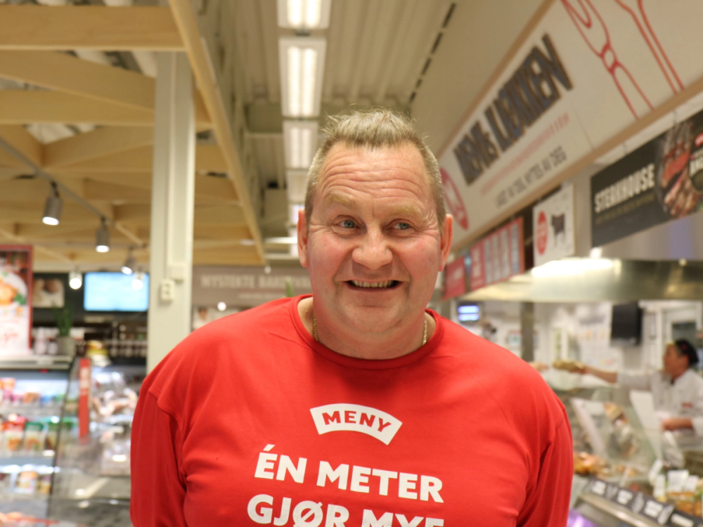 En person med kort hår i en butikk, iført en rød T-skjorte med teksten 'EN METER GJØR MEG', og butikkhyller i bakgrunnen.