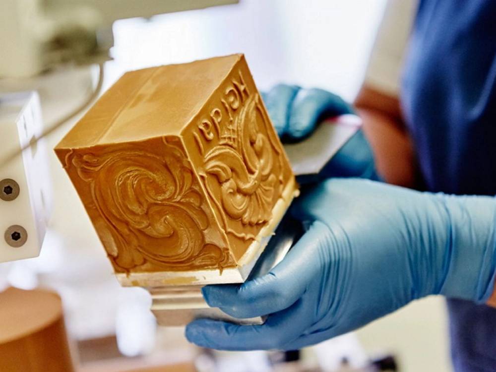 Person i blå hansker holder en stor blokk med brunost fra Heidal Ysteri, med en tydelig innpreget logo på siden.