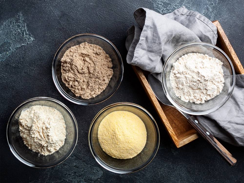 Ulike typer glutenfritt mel i små glass- og skålformer på en mørk bakgrunn.