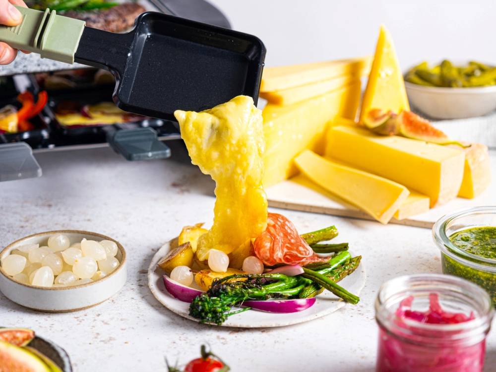 Raclette-ost smeltes over grønnsaker og serveres på en tallerken, med flere osteblokker og tilbehør i bakgrunnen.