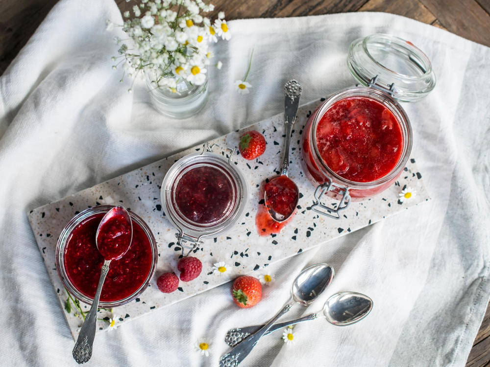 Ferske bær og syltetøy på et hvitt kjøkkenbord med skjeer og blomster i bakgrunnen