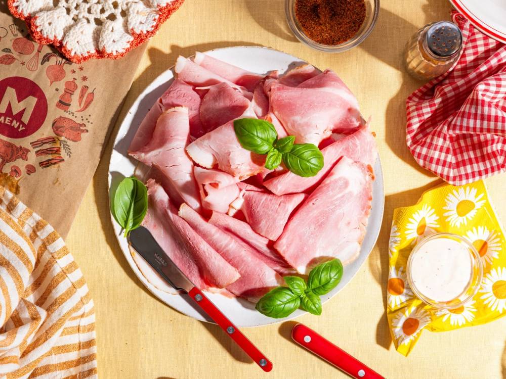 Skiver av fersk skinke med basilikum på en tallerken, omgitt av ulike matvarer og krydder på et kjøkkenbord.