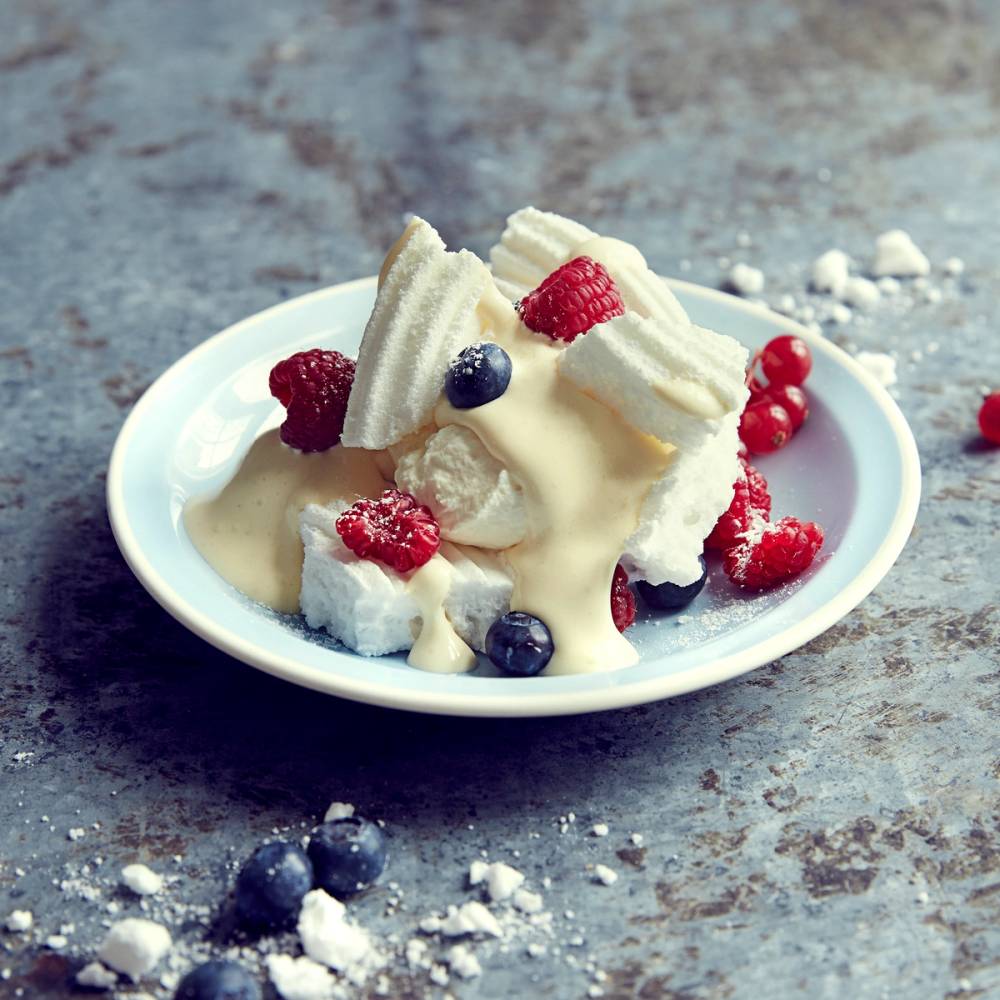 Pavlova med krem og ferske bær på en hvit tallerken, plassert på en rustikk overflate.