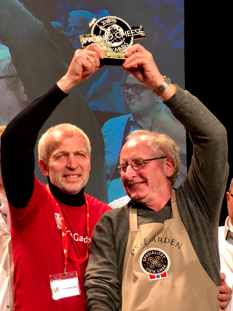 To personer holder opp en premie eller pokal med teksten 'World Cheese' under en seremoni eller konkurranse, med flere mennesker i bakgrunnen.
