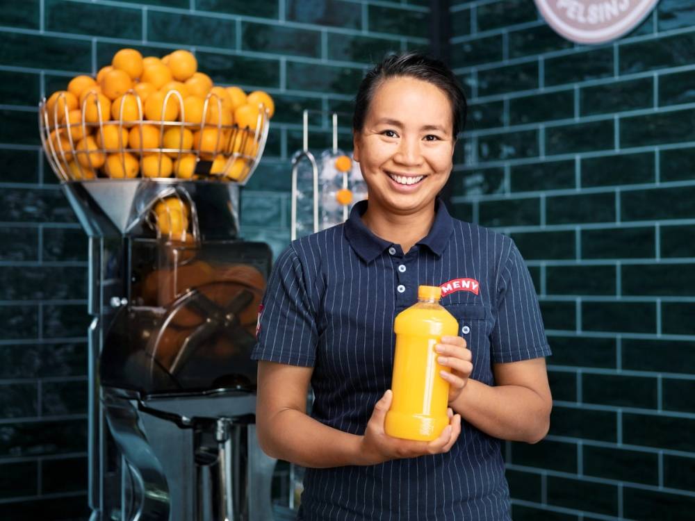 En person i en blå stripete skjorte som holder en gul flaske, med en frukt- og sjokoladeautomat i bakgrunnen, på Røa-menyen.
