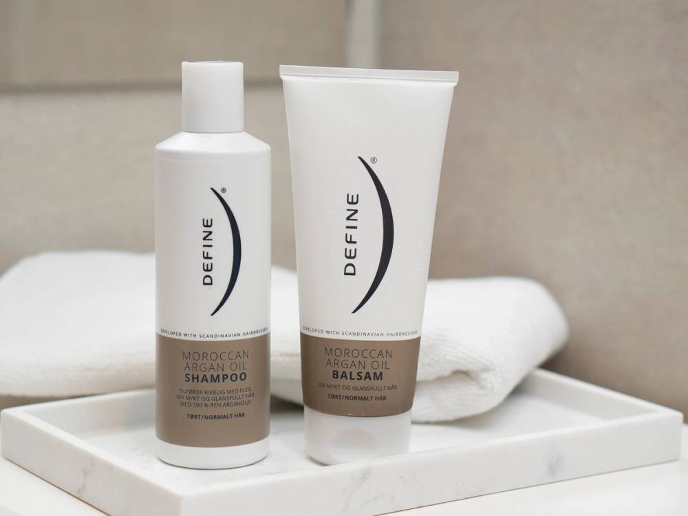 To hårpleieprodukter, sjampo og balsam, står på en hvit overflate med en hvit håndkle i bakgrunnen.