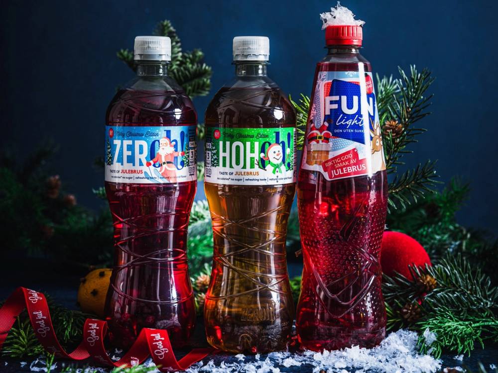 Tre flasker med julebrus står på en borddekorasjon med granbar og julepynt, mot en blå bakgrunn.