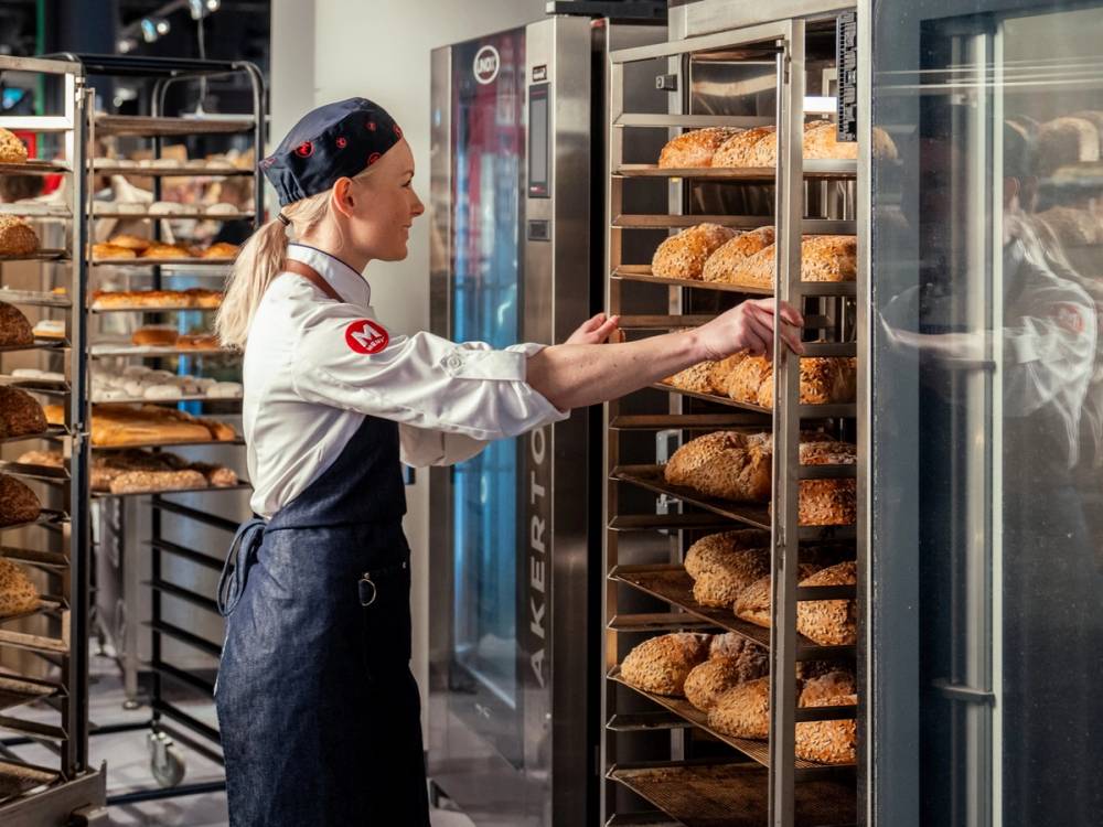 En person i bakeriutstyr tar ut brød fra en ovn eller bakeriovn, med flere brød på rister i bakgrunnen.