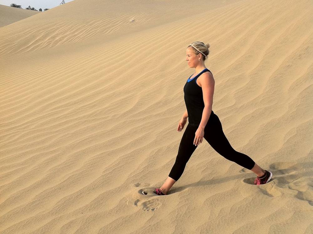 En person går oppover en sanddyne i ørkenen eller på en sandstrand, iført svart treningstøy og caps.