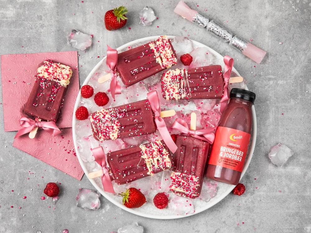 Fargerike sjokoladebarer med rosa og hvite striper, omgitt av friske jordbær og iskuber på en grå bakgrunn, med en liten flaske med drikke ved siden av.