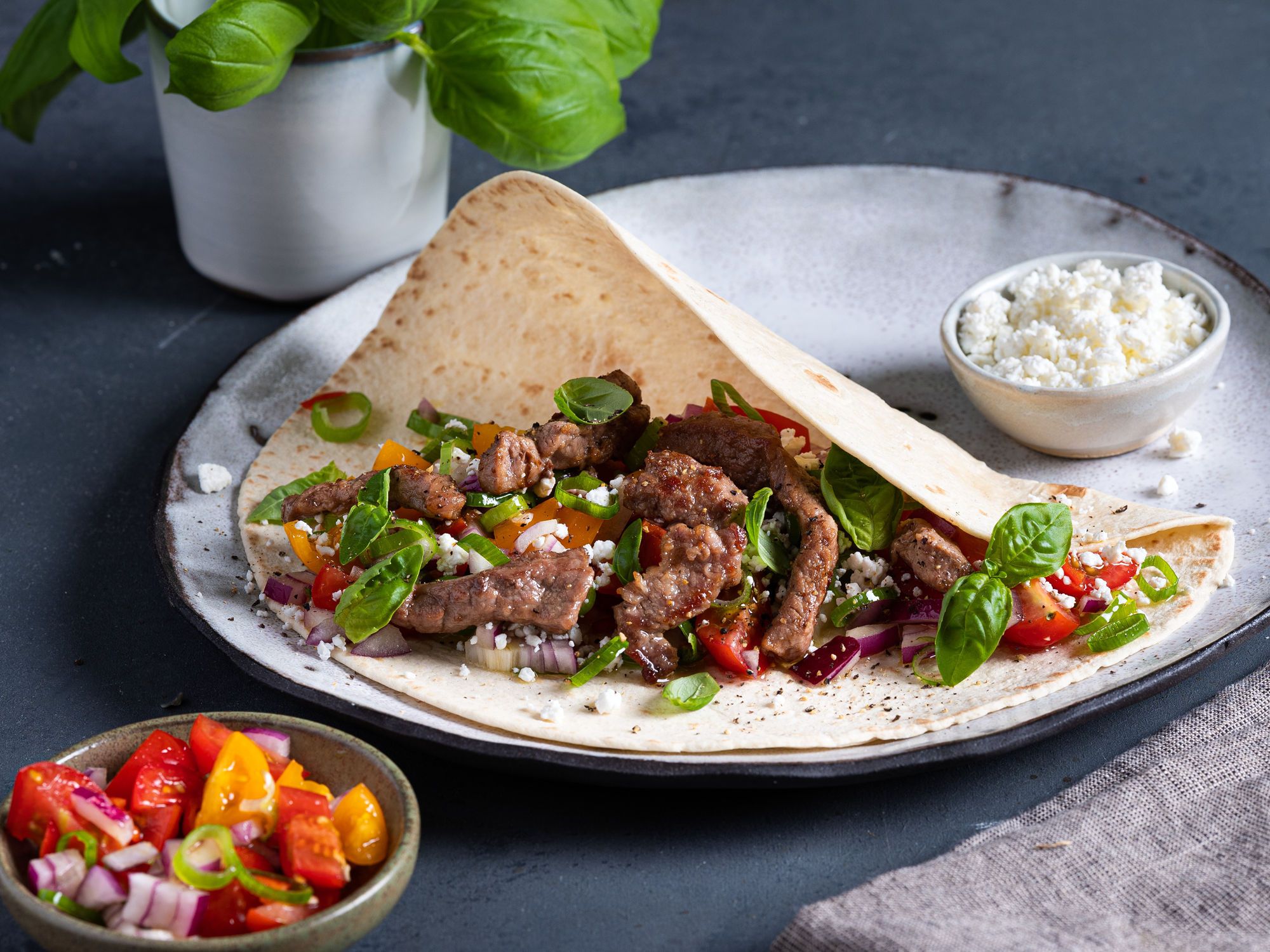Strimler av fersk lammelår i en tortilla med grønnsaker og ost, servert på en tallerken med en liten skål med ost og en annen med tomatsalat ved siden av.