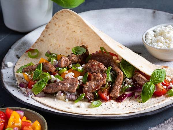 Strimler av fersk lammelår i en tortilla med grønnsaker og ost, servert på en tallerken med en liten skål med ost og en annen med tomatsalat ved siden av.