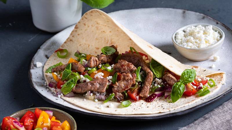 Strimler av fersk lammelår i en tortilla med grønnsaker og ost, servert på en tallerken med en liten skål med ost og en annen med tomatsalat ved siden av.