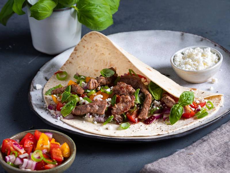 Strimler av fersk lammelår i en tortilla med grønnsaker og ost, servert på en tallerken med en liten skål med ost og en annen med tomatsalat ved siden av.